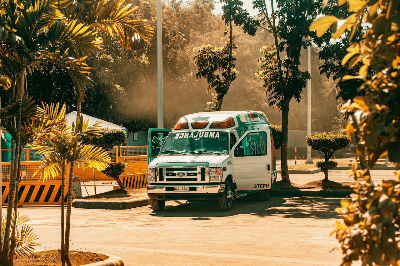 A parked ambulance in a sunny outdoor space, surrounded by trees and urban elements.